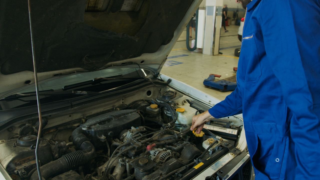 About Mechanic in blue coveralls inspecting an open car engine in an automotive workshop.